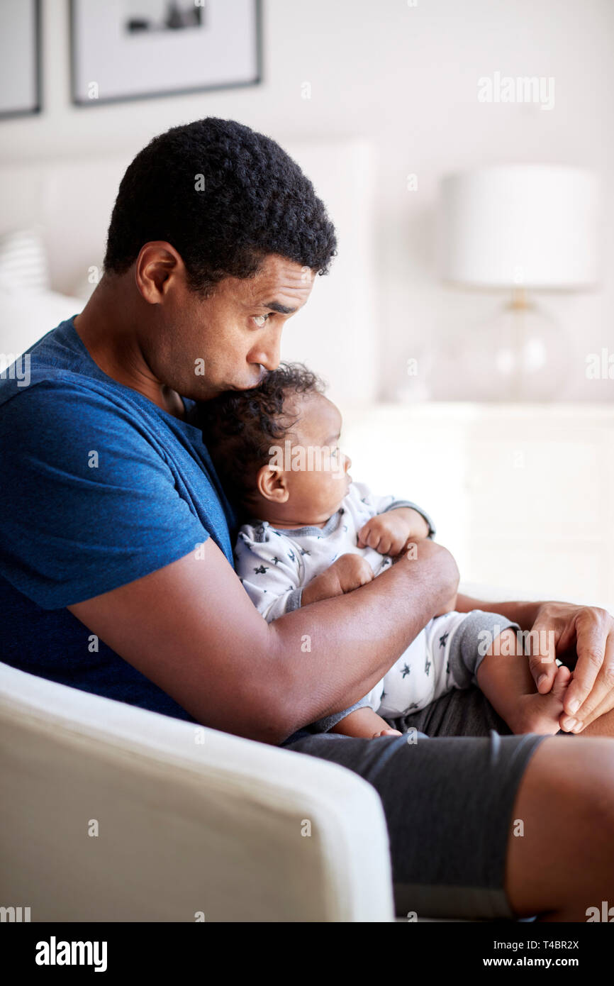 Young adult father sitting in an armchair holding his three month old baby son and kissing his head, side view, close up Stock Photo