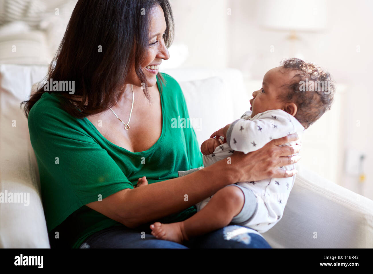 Middle aged mixed race grandmother sitting in an armchair holding her three month old grandson and smiling, focus on foreground Stock Photo