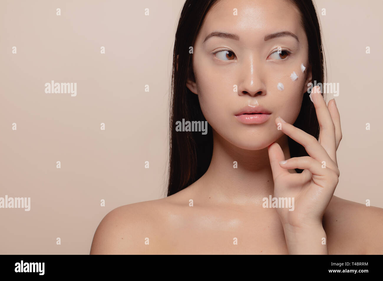 Close up of a beautiful young woman applying moisturizer to her face. Asian female model putting cosmetic cream on her face and looking away. Stock Photo