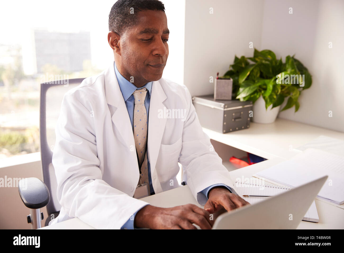 Male Doctor Wearing White Coat In Office Sitting At Desk Working On Laptop Stock Photo