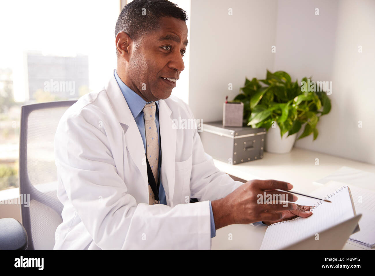 Male Doctor Wearing White Coat In Office Sitting At Desk Working On Laptop Stock Photo