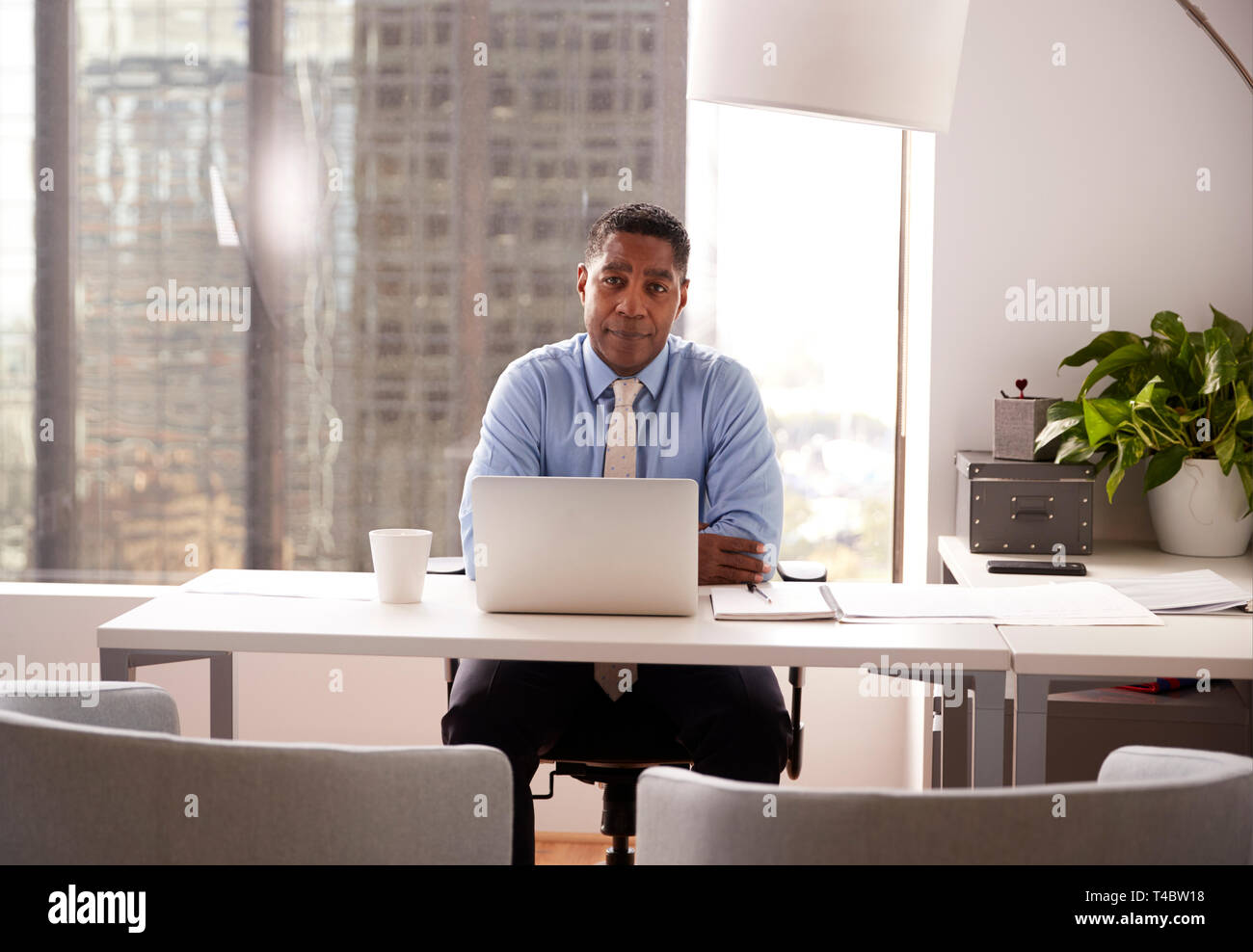 Portrait Of Male Financial Advisor In Modern Office Sitting At Desk Working On Laptop Stock Photo