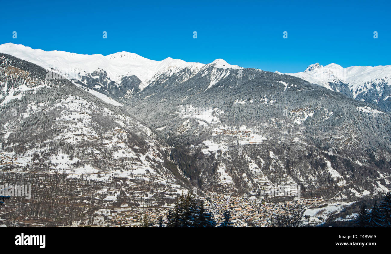 Panoramic view down snow covered valley in alpine mountain range with conifer pine trees on blue sky background Stock Photo