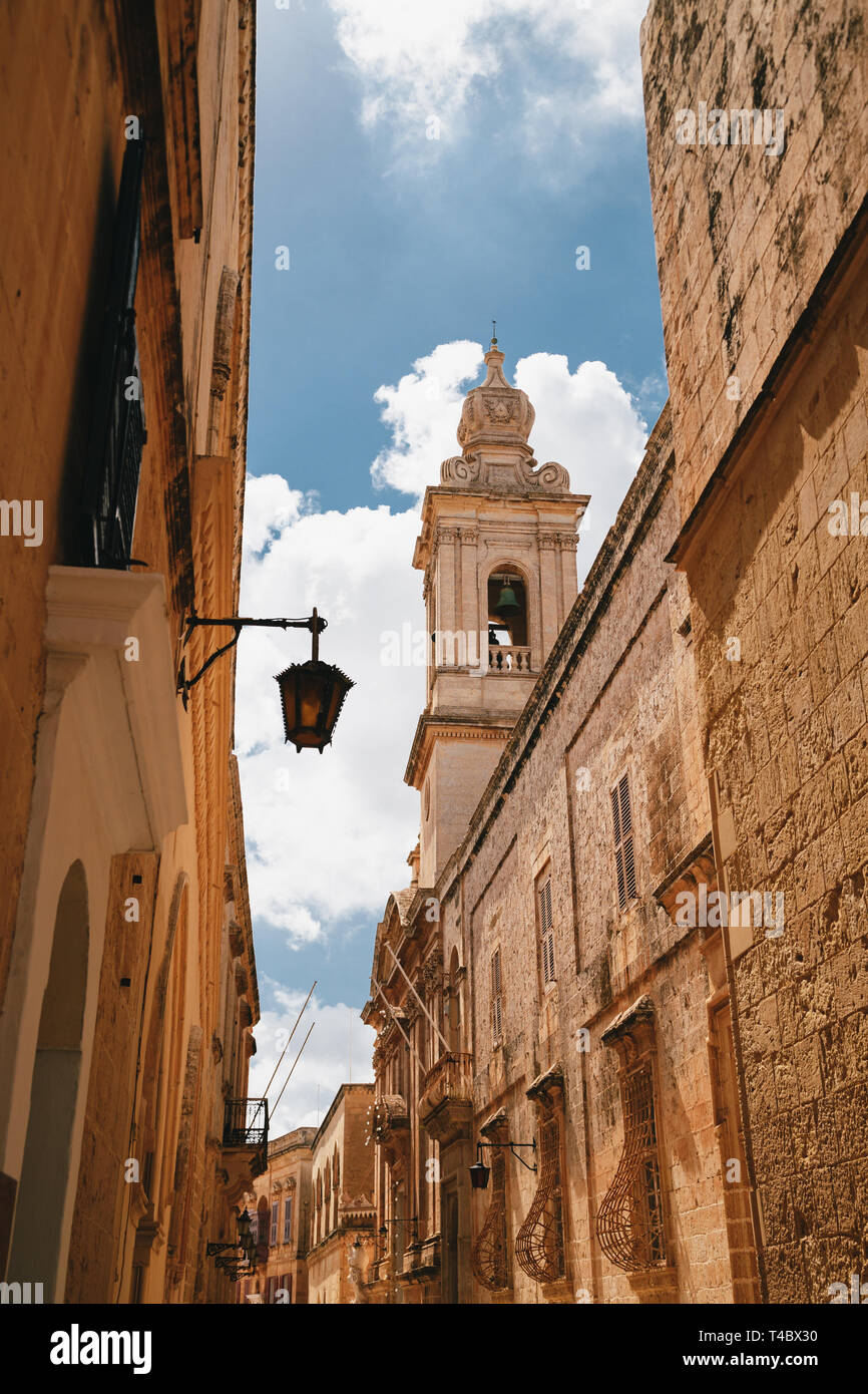 Beautiful view of narrow medieval street in Mdina with Carmelite Church Bell Tower an blue sky with clouds at the background. Ancient capital of Malta Stock Photo