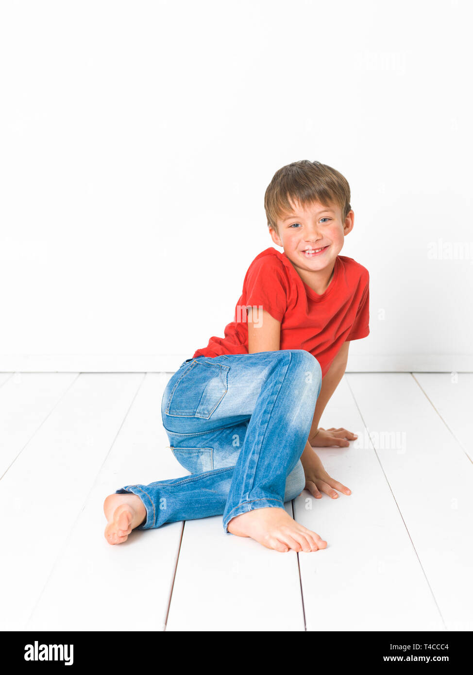 cute and blond boy with red shirt and blue jeans is posing on white wooden  floor in front of white background Stock Photo - Alamy