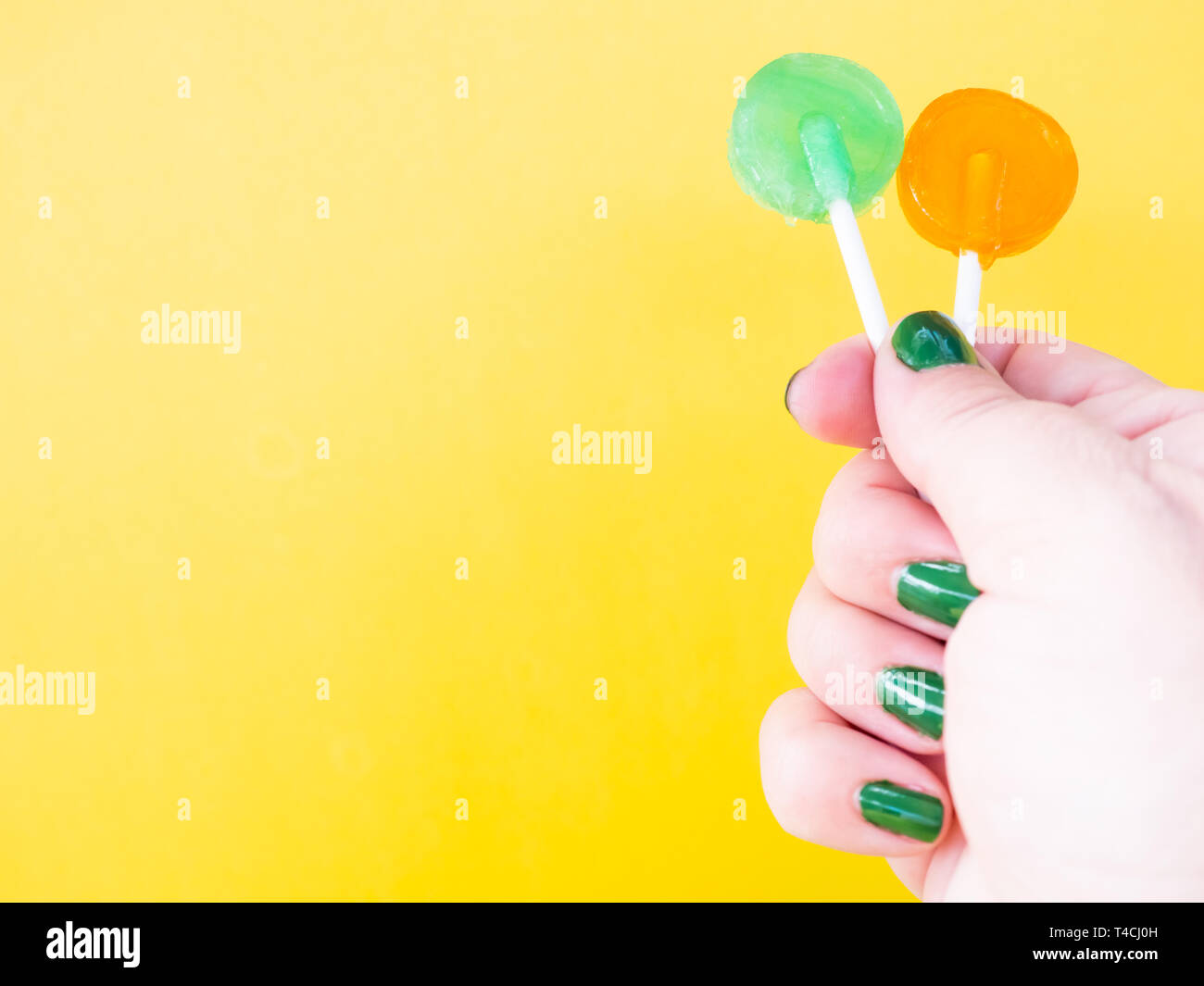 A woman with green painted nails with two different lollipop in her hand and yellow background Stock Photo