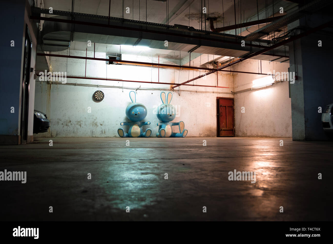 Two huge blue bunny figurines laying on the ground in a dark, spooky underground garage, with grungy water pipes on the ceiling and a rusty exit door. Stock Photo