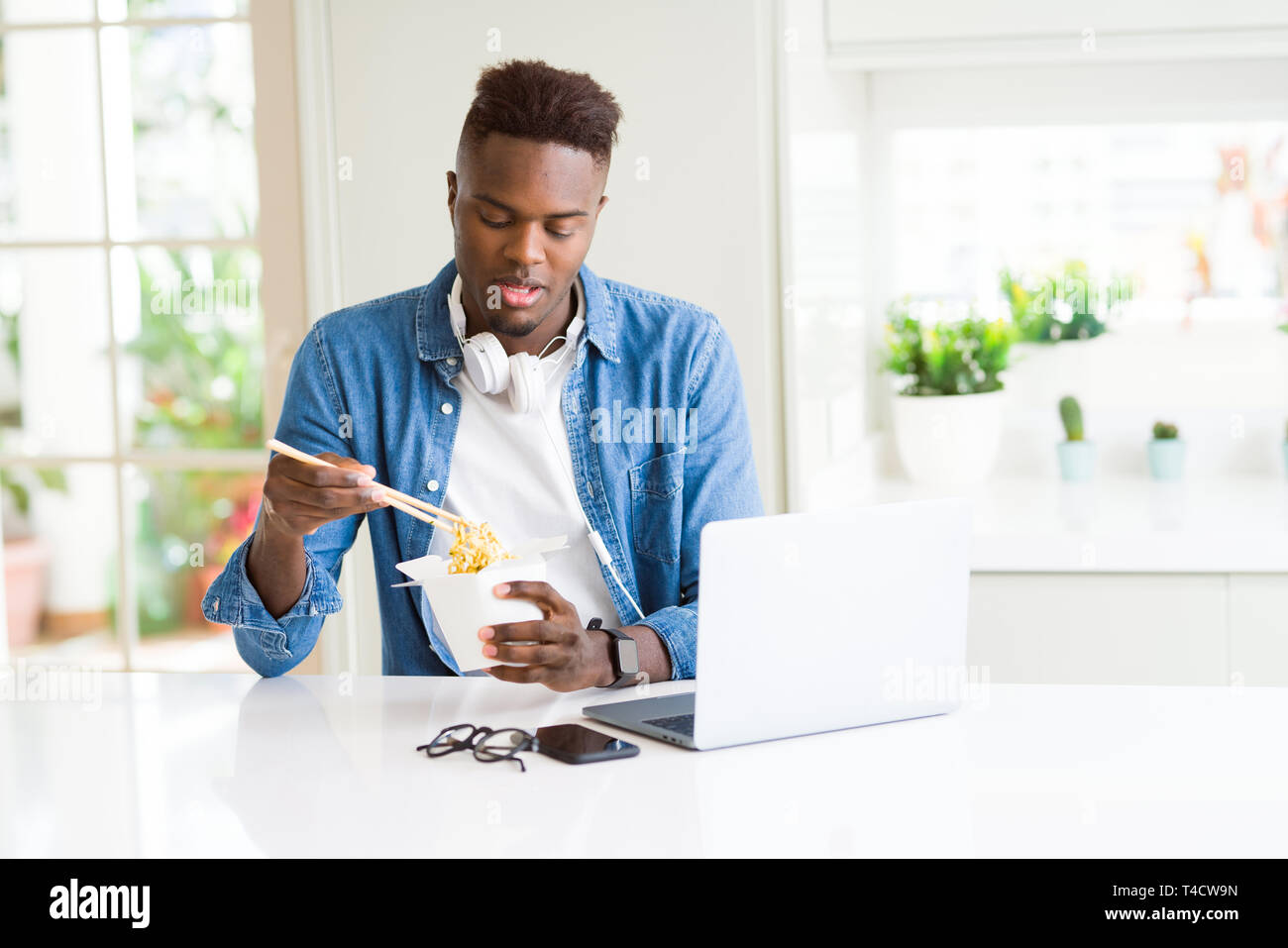Handsome young african business man eating delivery asian food and working using computer, enjoying noodles smiling Stock Photo