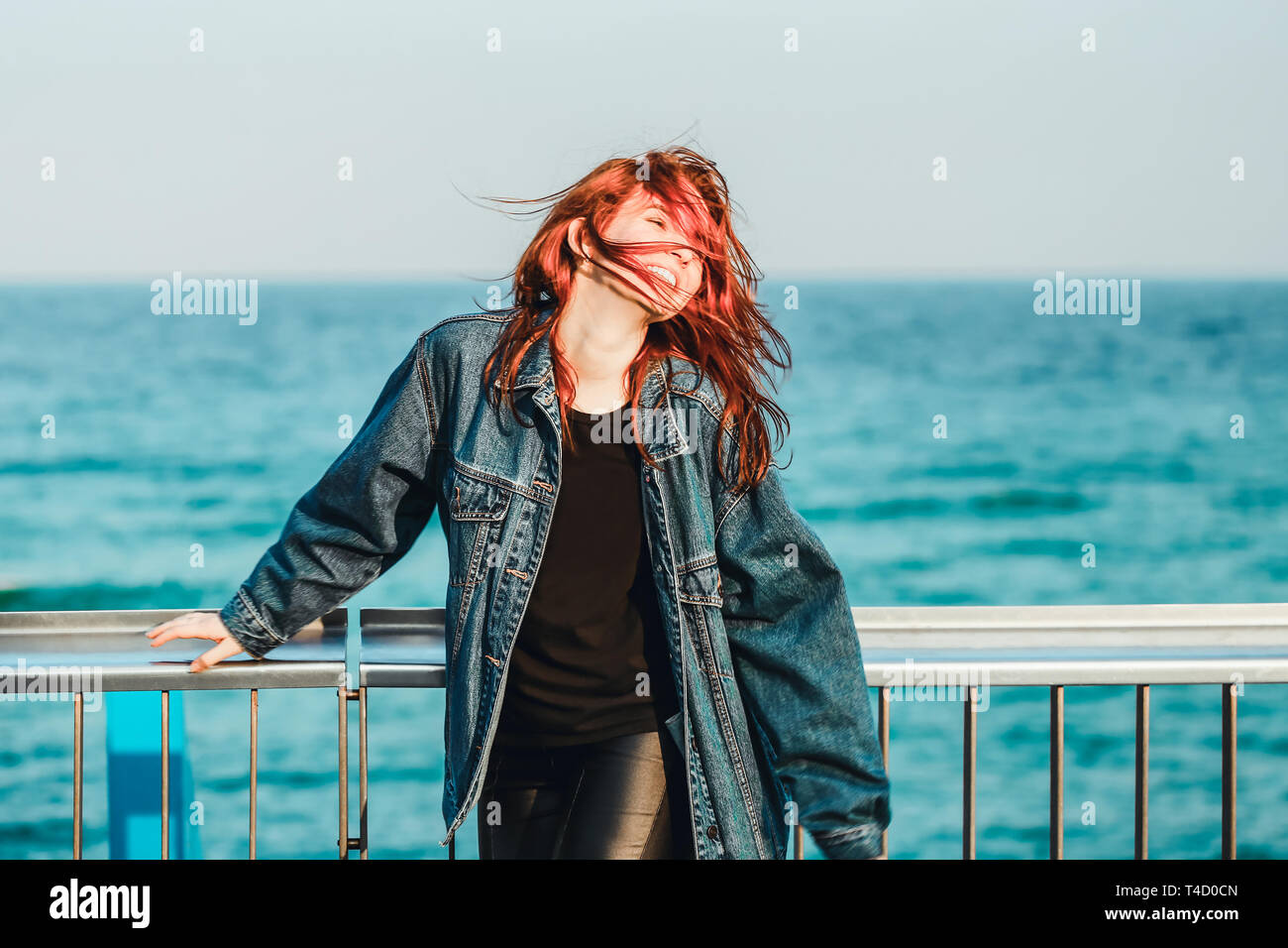 Young nice red-haired girl with jeans jacket walking next to sea water and feeling happy and free Stock Photo