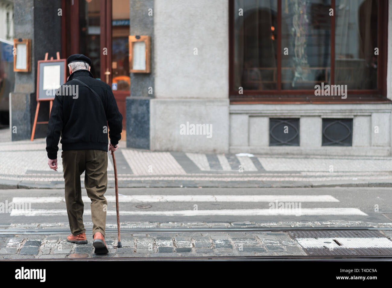 An elderly male with a walking stick slowly walks across a crosswalk in the middle of a city alone Stock Photo