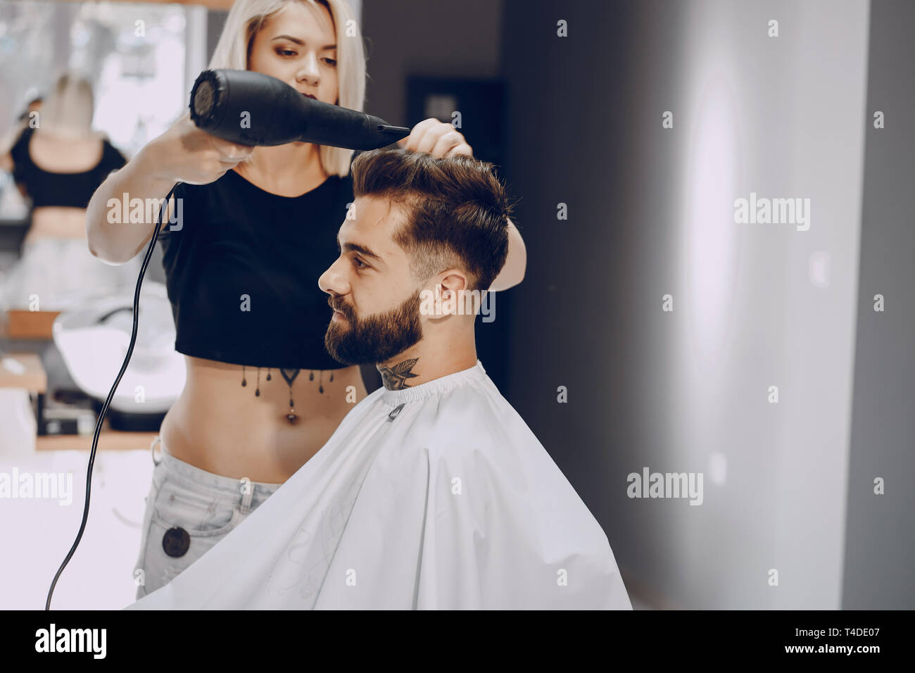 handsome young bearded guy sitting in an armchair in a beauty salon and the girl around him dry his hair Stock Photo