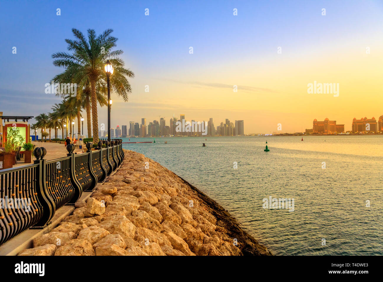 Palm trees along marina walkway in Porto Arabia at the Pearl-Qatar, Doha, with skyscrapers of West Bay skyline at sunset sky. Scenic sunset landscape Stock Photo