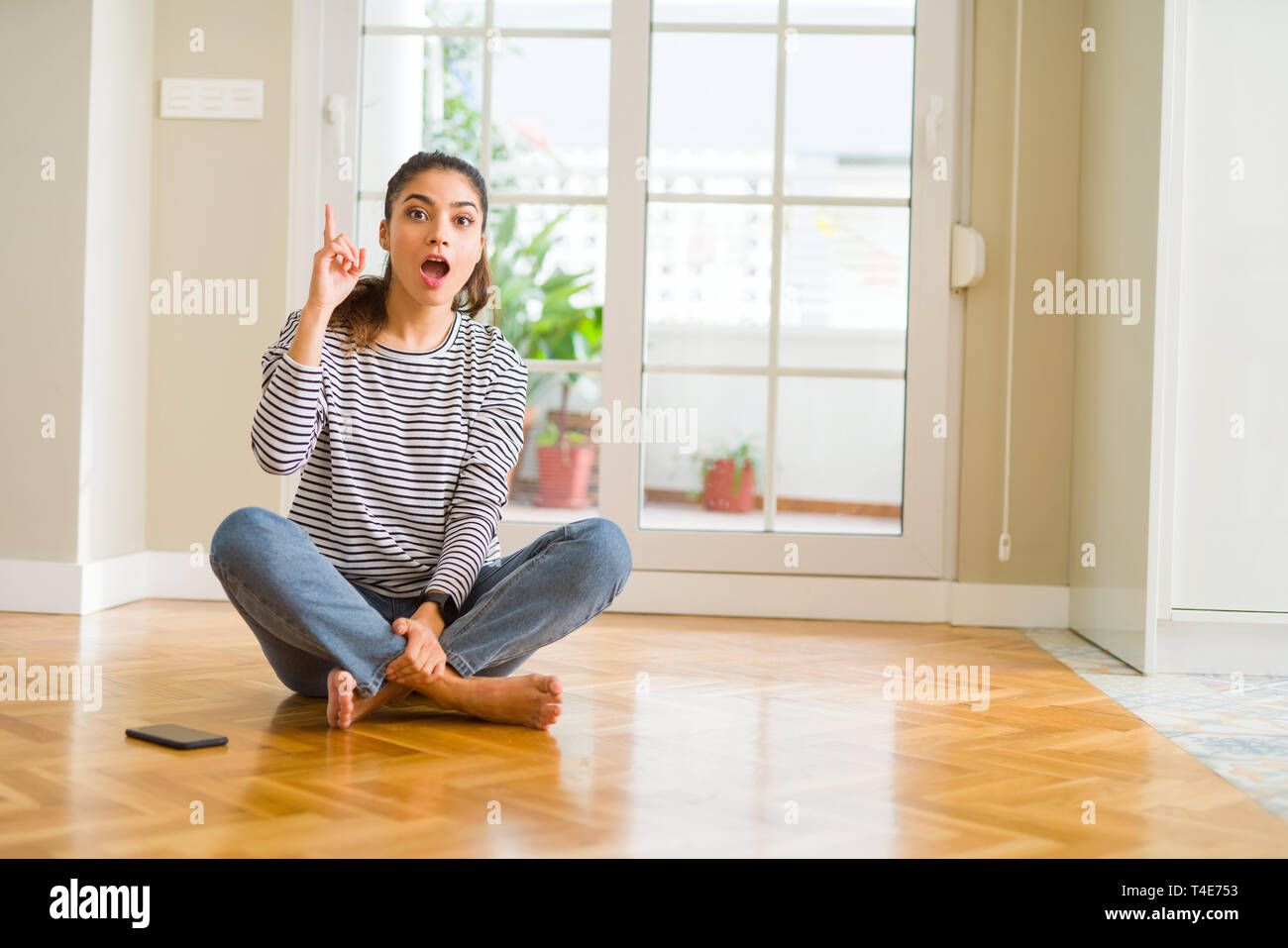 Young beautiful woman sitting on the floor at home pointing finger up ...