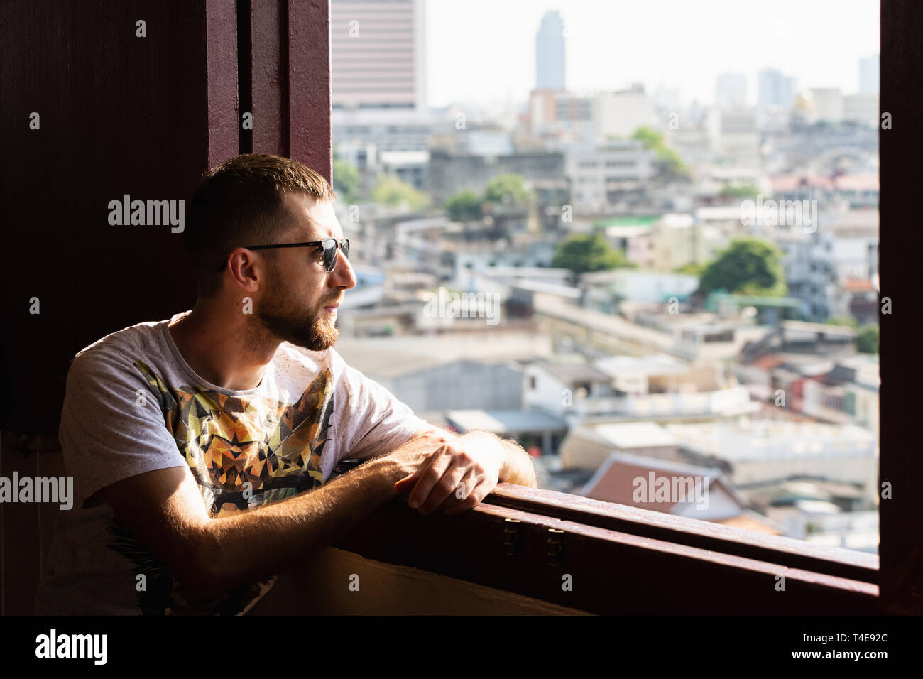 Bearded young man with sunglasses looking out of Golden Mount Wat Saket temple window over Bangkok, Thailand. Stock Photo