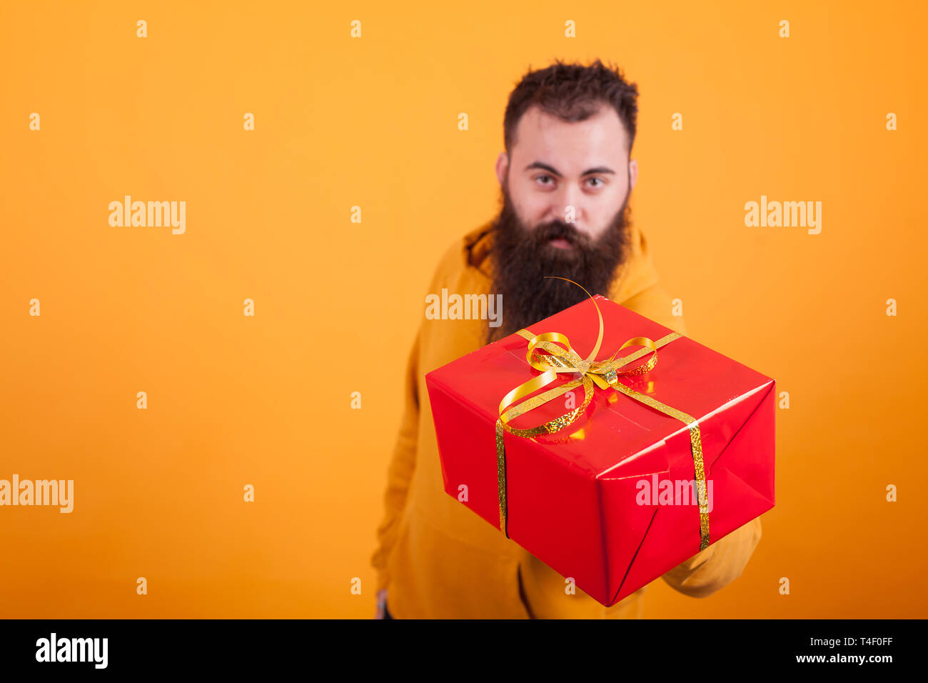 Attractive bearded man wearing yellow hoodie shoing red gift box over yellow background. Giving red gift box. Stock Photo