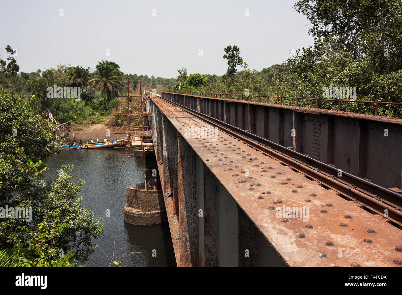 Rail & port operations for managing & transporting iron ore. Overhaul of river rail bridge near Port Loko with village river boat taxi passing beneath Stock Photo