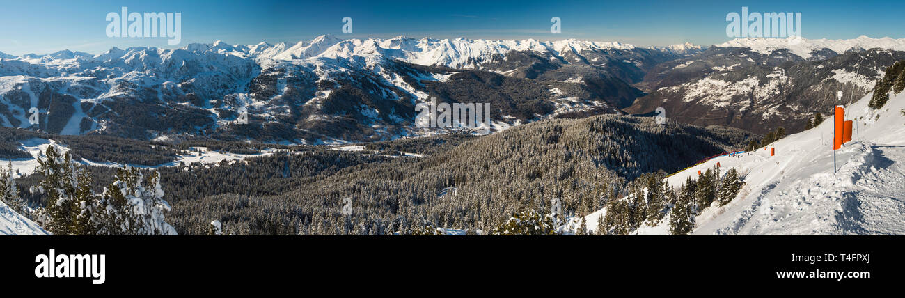 Panoramic view down snow covered valley in alpine mountain range with conifer pine trees on blue sky background Stock Photo