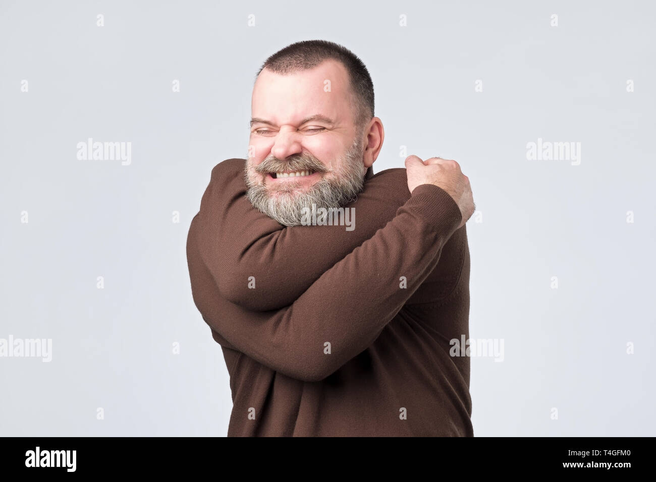 Funny playful european mature guy with beard cuddling himself and smiling from pleasure, closing eyes and dreaming about finding soulmate Stock Photo