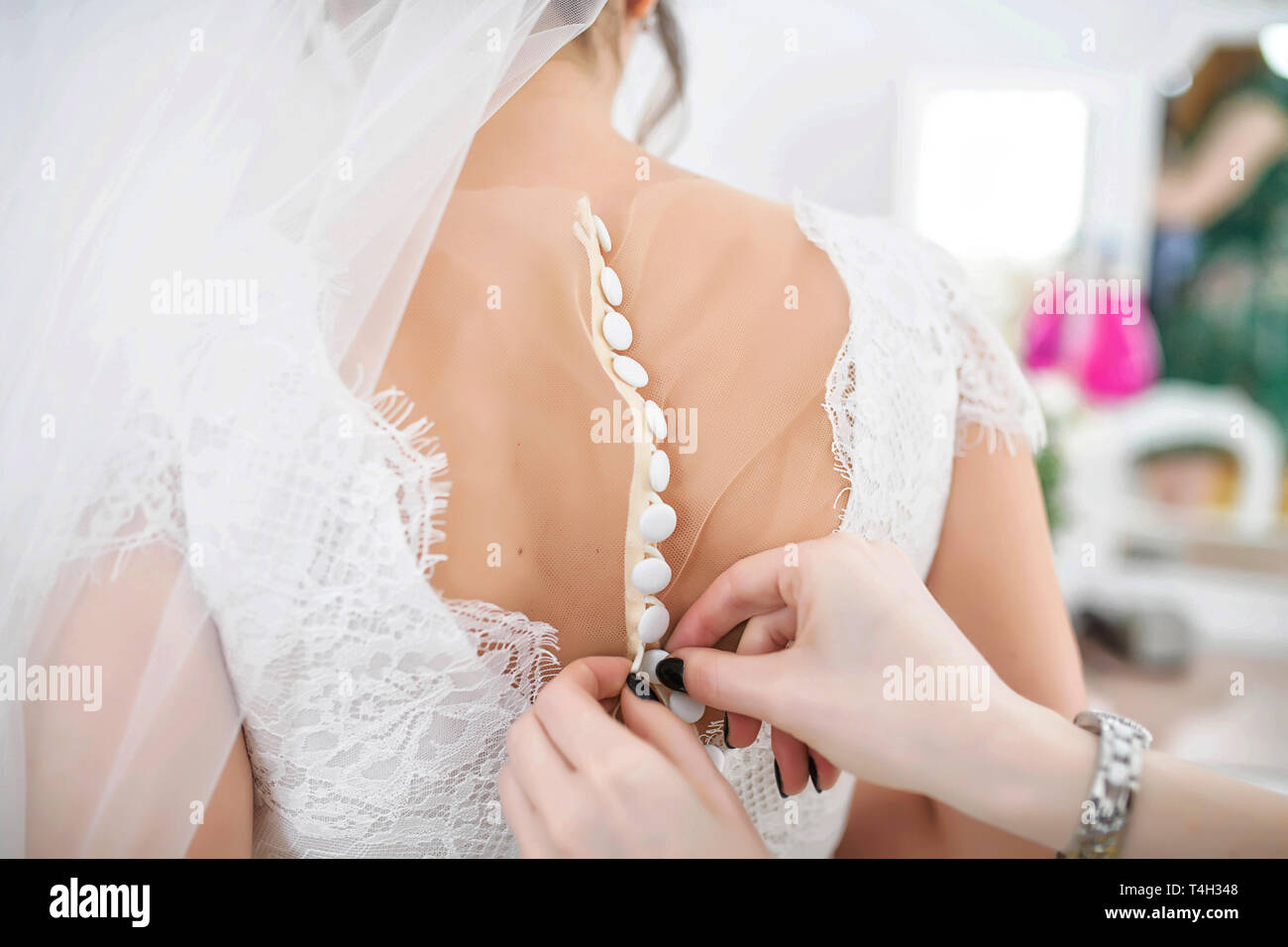 Bridesmaid or maid of honor helping the  bride buttoning up the lacy embroidered white dress at the back, in preparation for the wedding ceremony Stock Photo