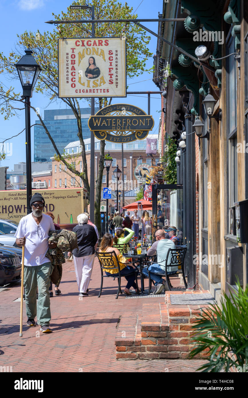 Fells Point, Baltimore, MD, USA -- April 13, 2019. A man with a cane walks along Thames St while others have breakfast at an outdoor cafe in Fells Poi Stock Photo