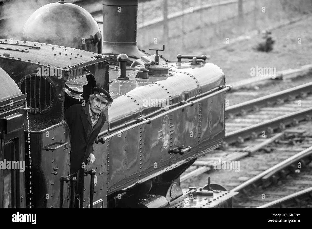 Black & white close up of vintage UK steam locomotive on railway track arriving at station; steam train crew driver in cab leaning out, looking up. Stock Photo