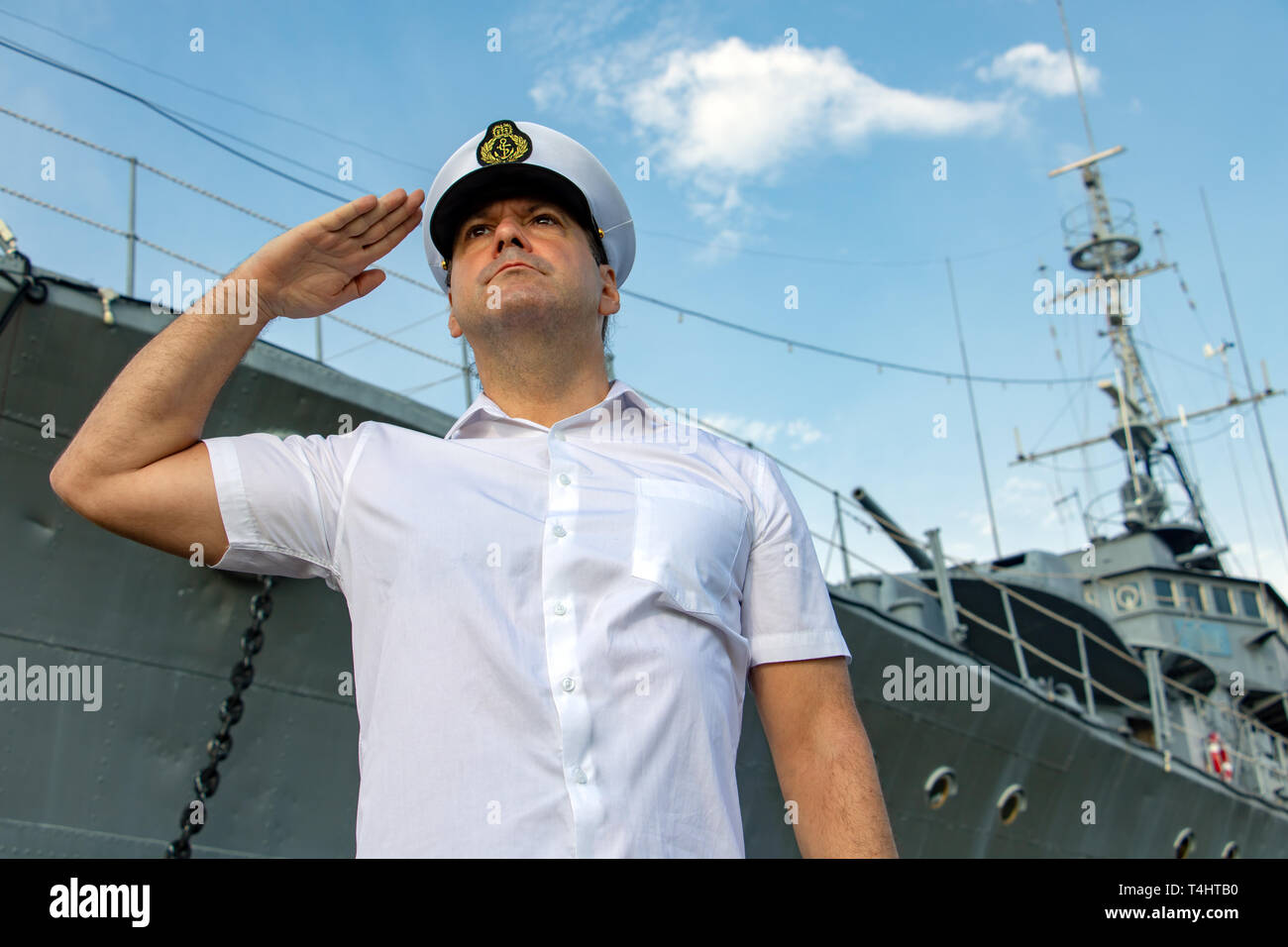 avy officer standing beside warship and do salute.The captain in white uniform stands under a battleship and saluting. Stock Photo