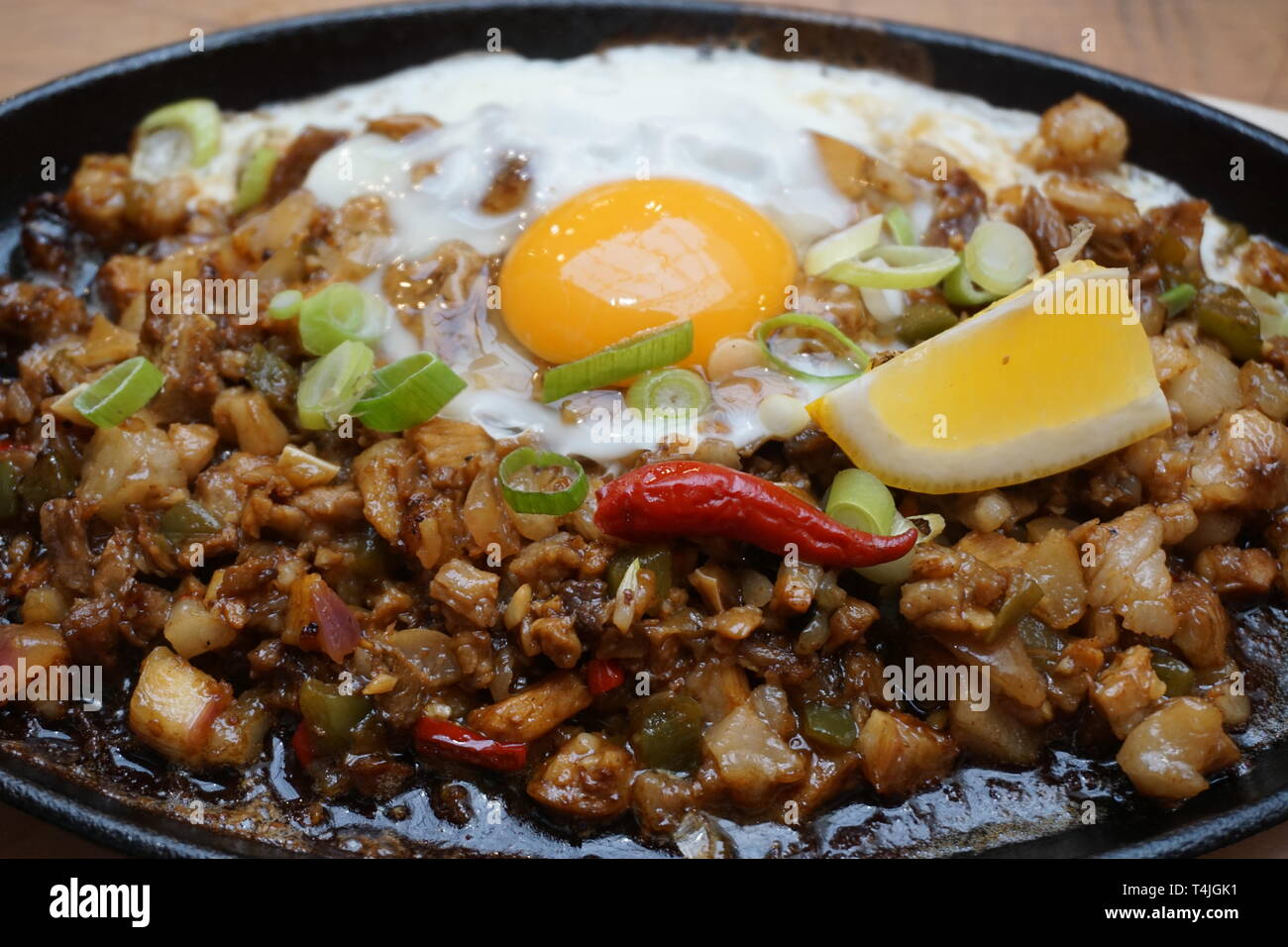 Delicious Pork sisig sizzling mince pork Filipino food with raw egg cooking hot on clay pot tray with lemon slice and fresh red chilies Asian cuisine Stock Photo