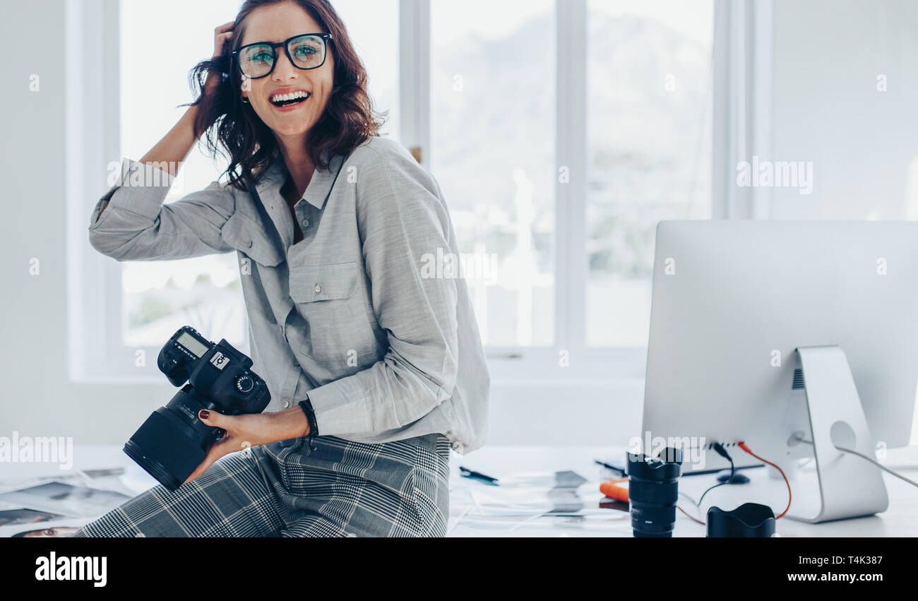 Smiling female photographer with a professional camera sitting on her desk. Woman with dslr camera in office looking away and smiling. Stock Photo