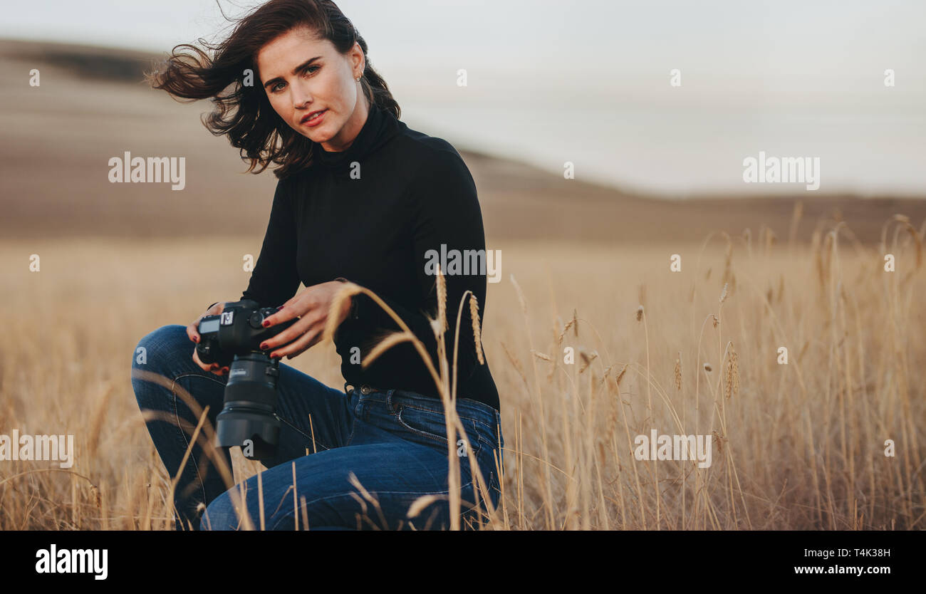 Woman photographer with camera crouching in a dry grass field. Woman doing landscape photography outdoors. Stock Photo