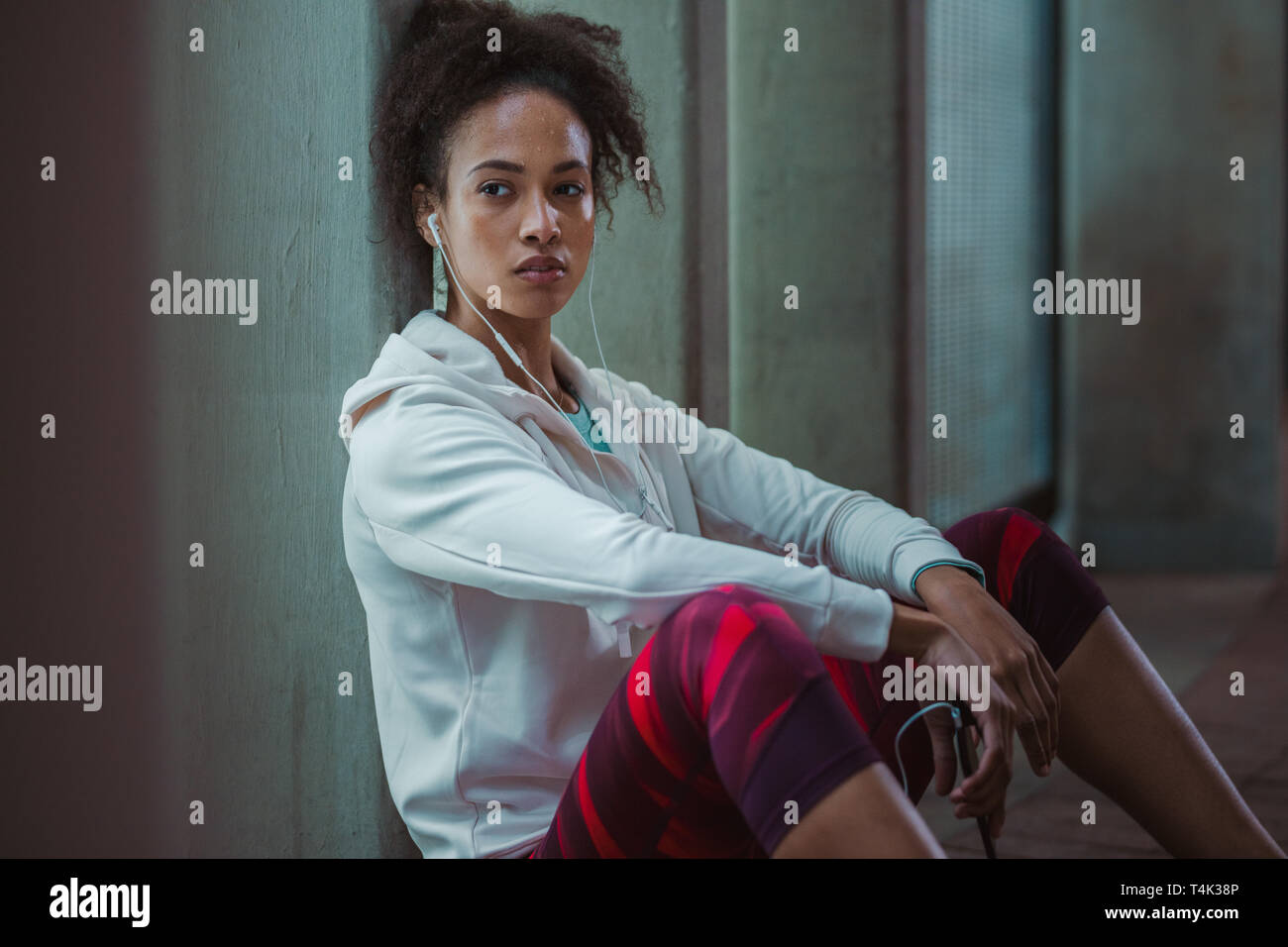 Woman sitting by the street leaning to a pillar and looking away. Urban runner resting after training session. Stock Photo