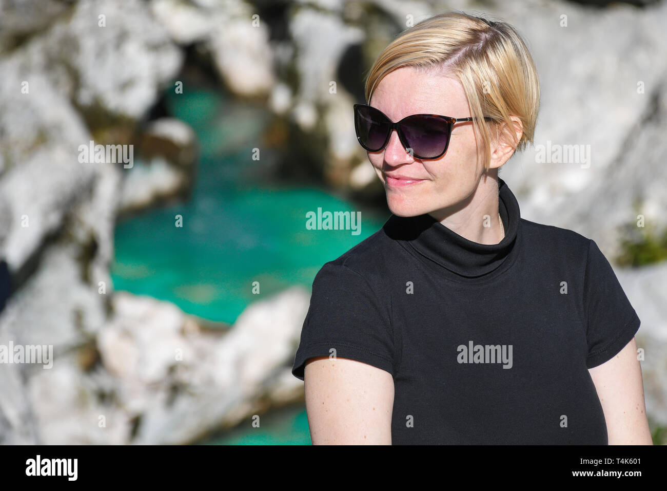 Happy smiling woman with black sunglasses and shirt posing next to a beautiful turquoise colored Soca river canyon in the background, Slovenia Stock Photo