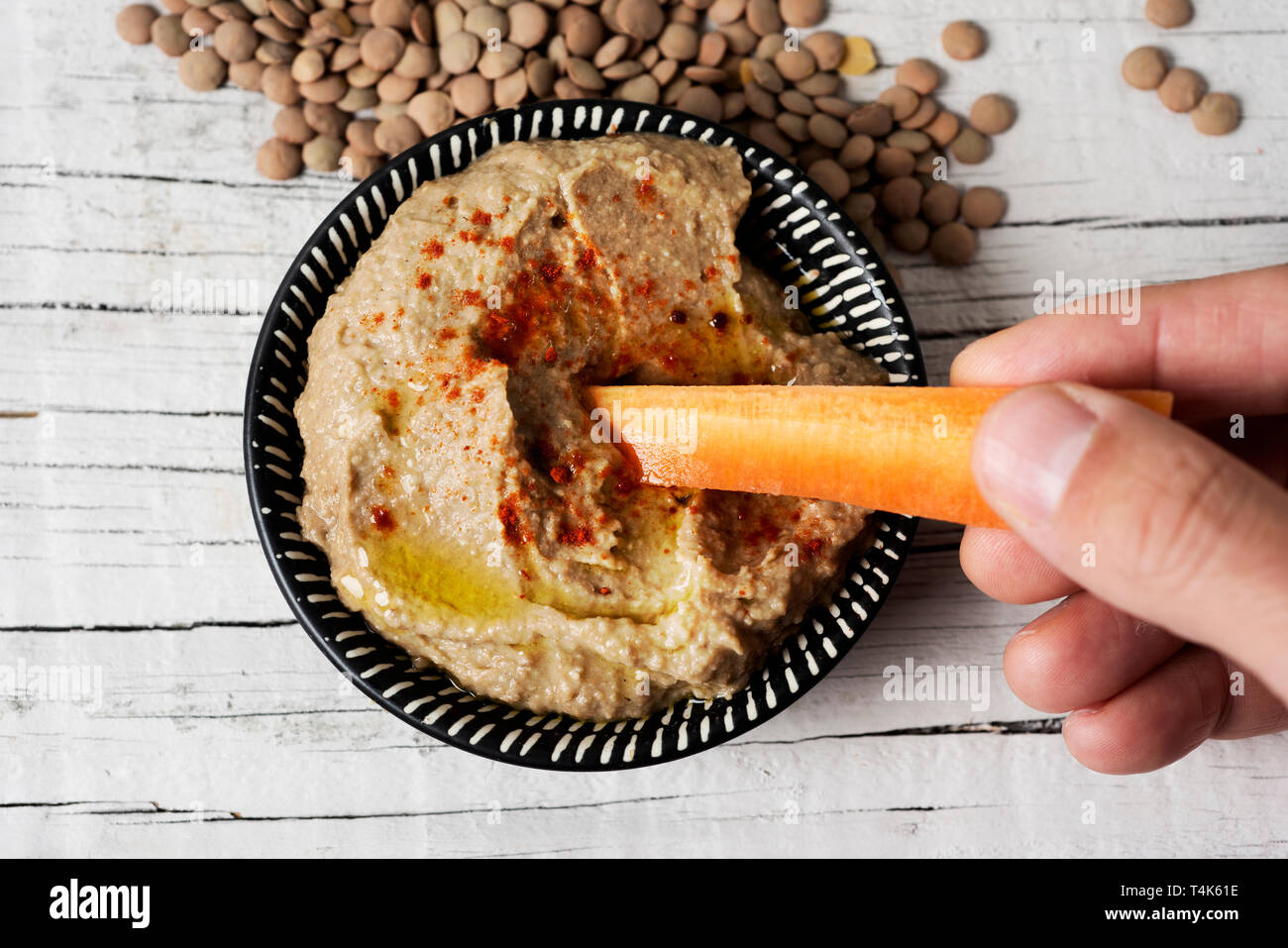 high angle view of a man dipping a strip of carrot in a homemade lentil hummus seasoned with paprika served in a green ceramic plate, on a white rusti Stock Photo