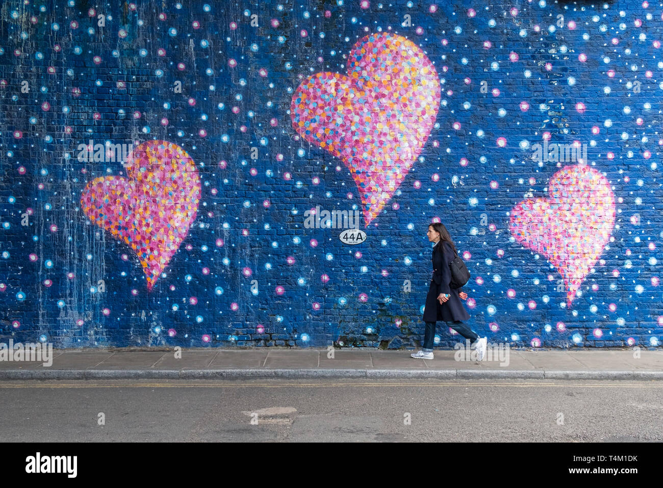 A woman walking along a street next to a wall covered in a painted mural of colourful bubbles and hearts. Stock Photo