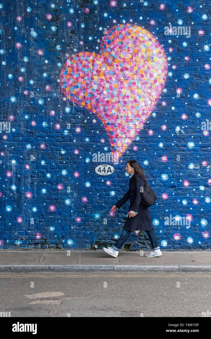 A woman walking along a street next to a wall covered in a painted mural of colourful bubbles and hearts. Stock Photo