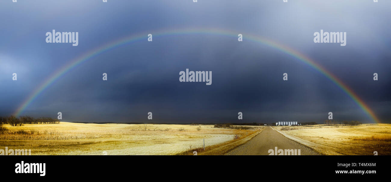 A full colorful rainbow over a gravel road between harvested golden fields Stock Photo