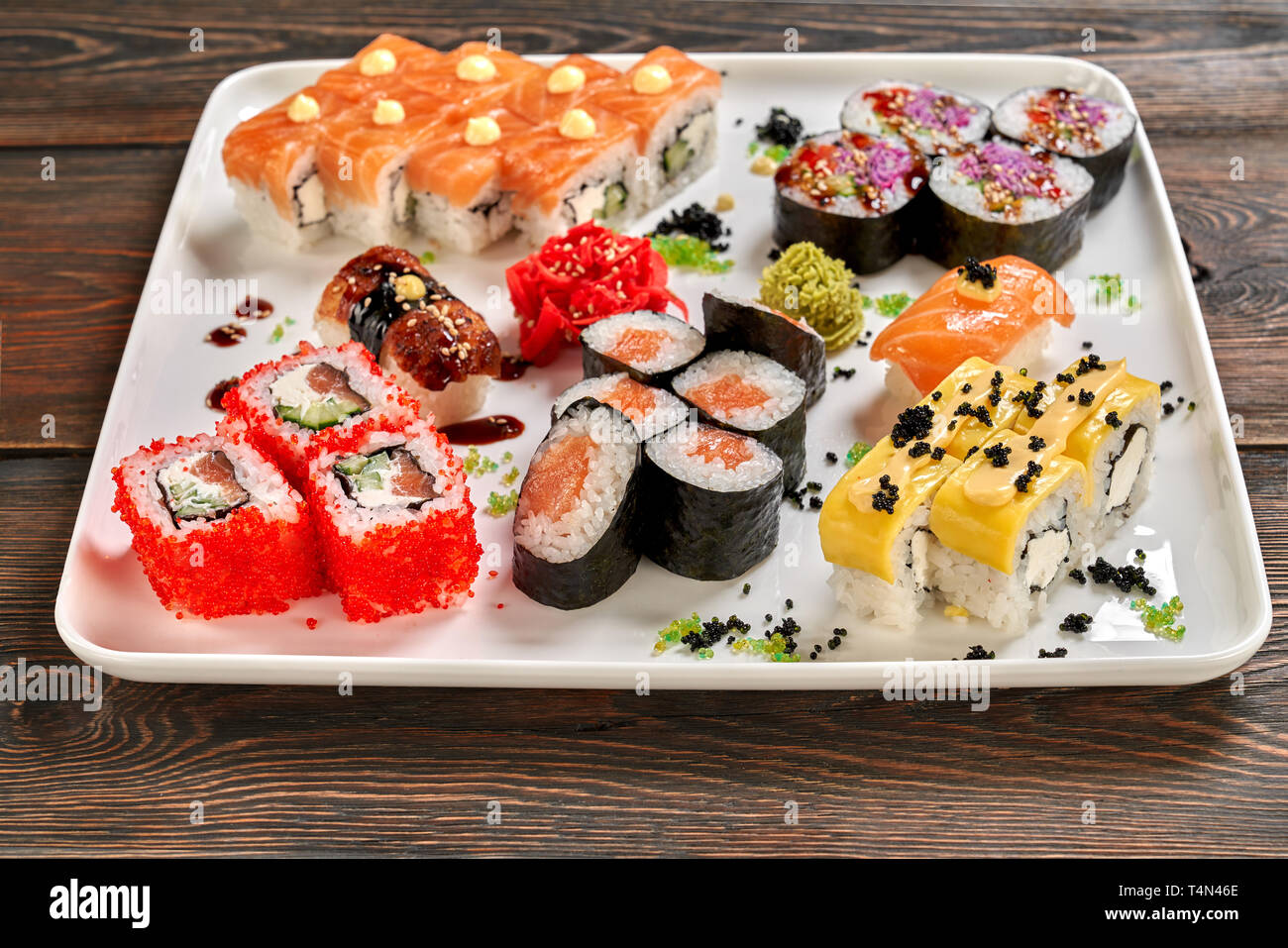 Different kinds of nigiri, maki and uramaki. Philadelphia, cheese uramaki, veggie rolls and nigiri served with wasabi and pickled ginger on white plate, wooden background. Stock Photo