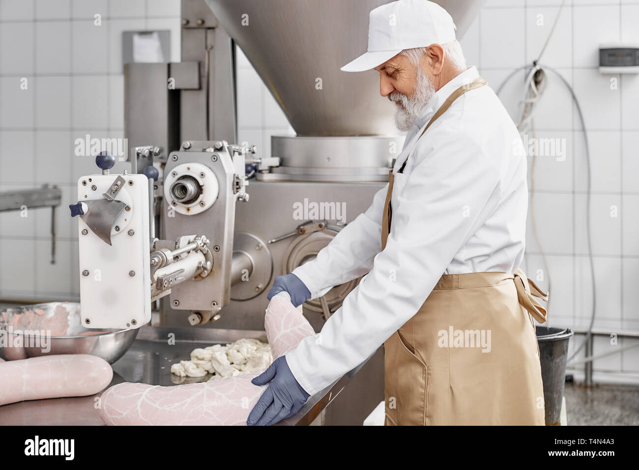Butcher man working with production of sausages, standing near equipment, holding sausage. Worker wearing in white uniform, brown apron, rubber gloves. Food industry. Stock Photo