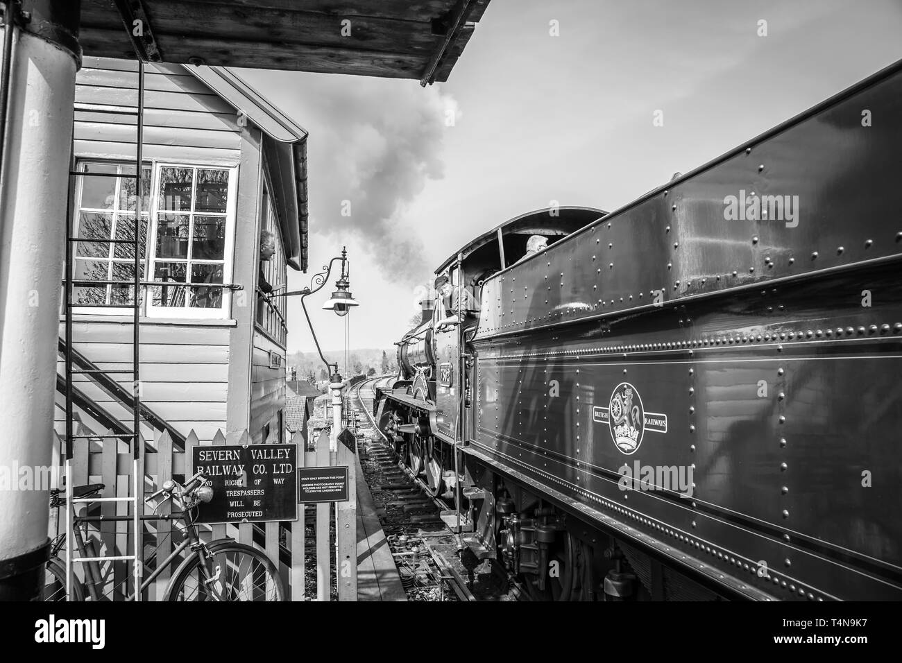 Black & white scene of railway signalman at vintage station looking out of signal box window greeting cab crew of puffing vintage steam train passing. Stock Photo