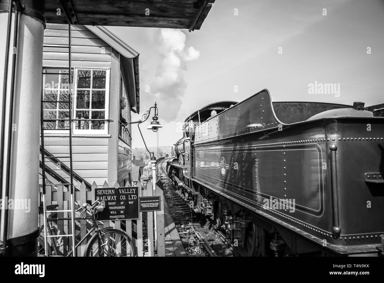 Black & white scene of railway signalman at vintage station looking out of signal box window greeting cab crew of puffing vintage steam train passing. Stock Photo