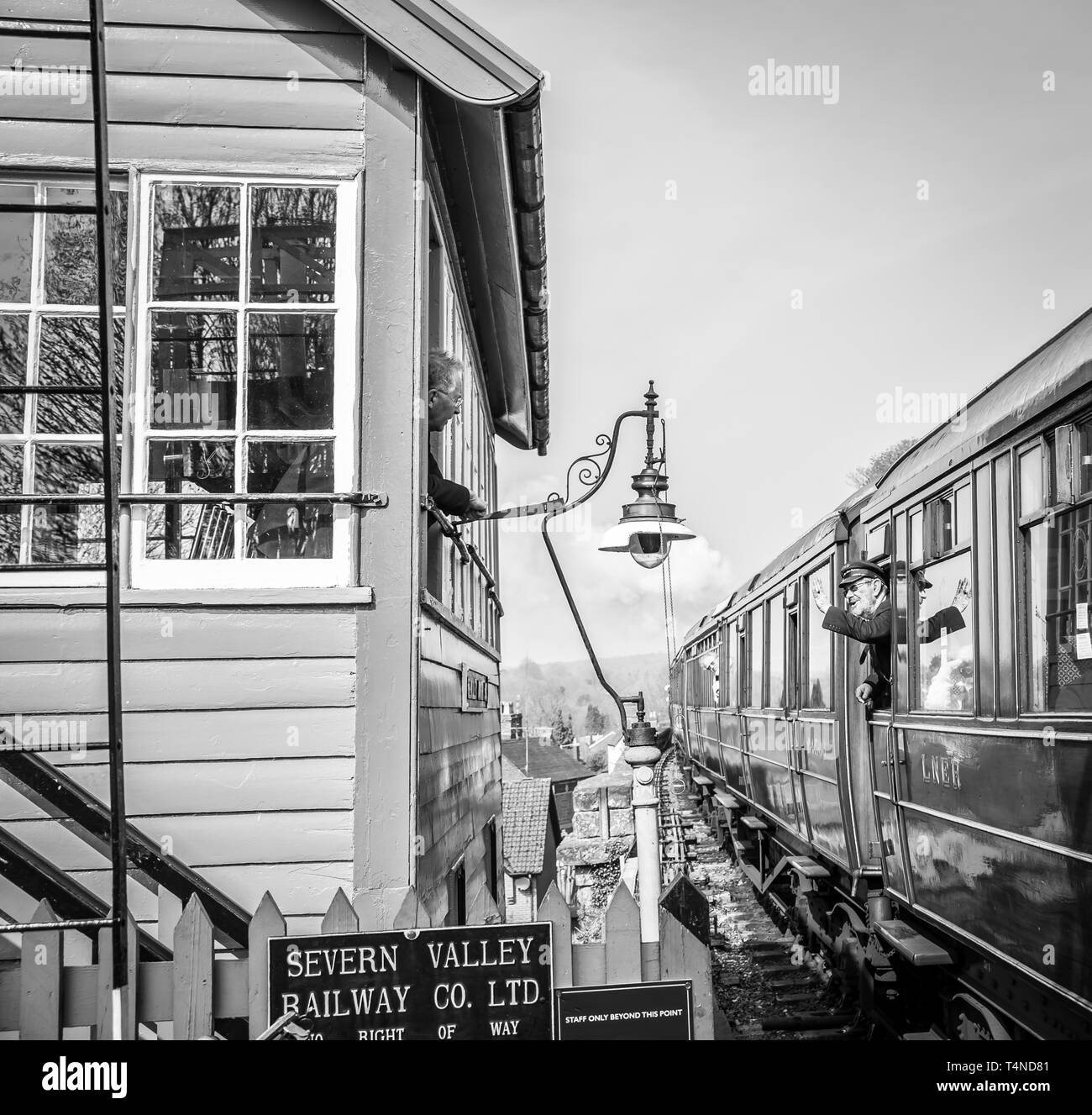 Railway signalman on SVR heritage line looks out window to greet train guard waving from inside vintage steam train passing his signal box. Stock Photo