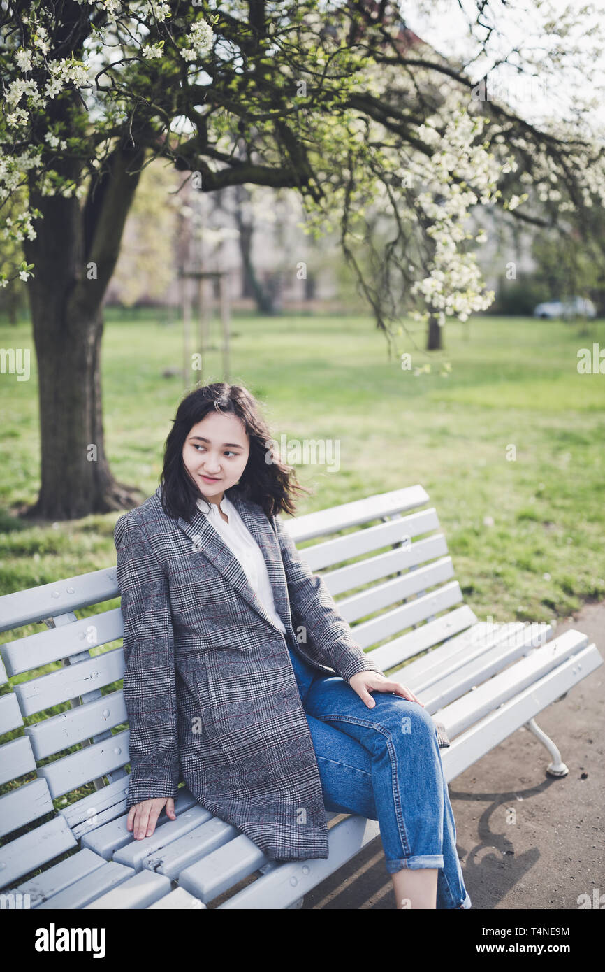 Happy asian young woman in a tweed coat and jeans sitting on a bench ...