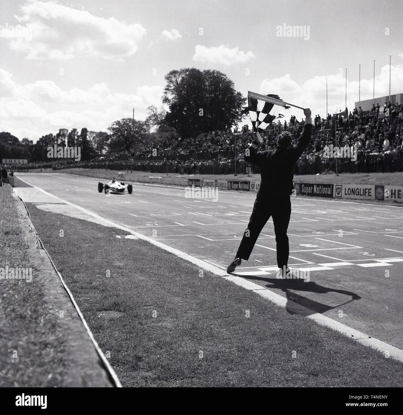 1960s, historical, motor racing at Crystal Palace race circuit in Crystal Palace Park in South London, England, UK. A race official by the finishing line of the track with the chequered flag raised as the winning car comes down the finishing straight in front of the crowd. Opened in 1927 for motorcycles, it was 1937 when 20 cars entered the first London Grand Prix. Later that year, the BBC broadacst the first ever televised motor racing from the circuit. The final meeting was in 1972 when it was felt that racing around a park at speeds of over 100 mph was not safe. Stock Photo