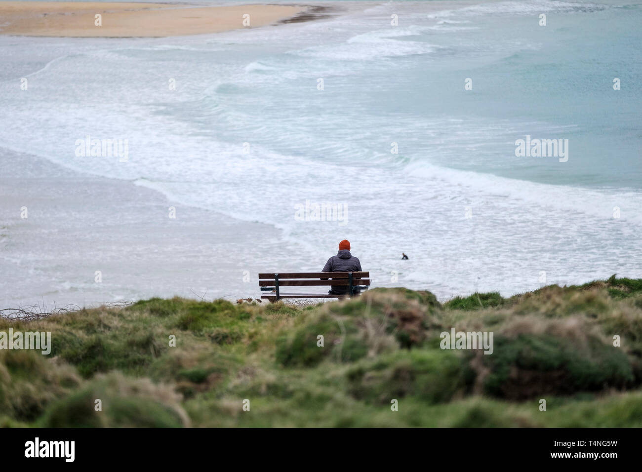 A man sitting alone on a bench overlooking a deserted Crantock Beach in Newquay in Cornwall. Stock Photo