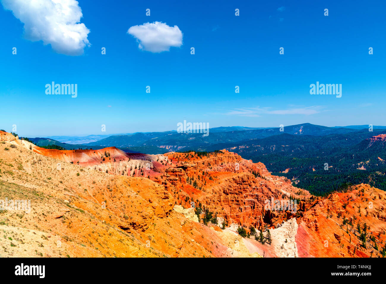 Yellow dirt makes way for orange and reddish rock formations in canyon with green mountains beyond under a bright blue sky. Stock Photo