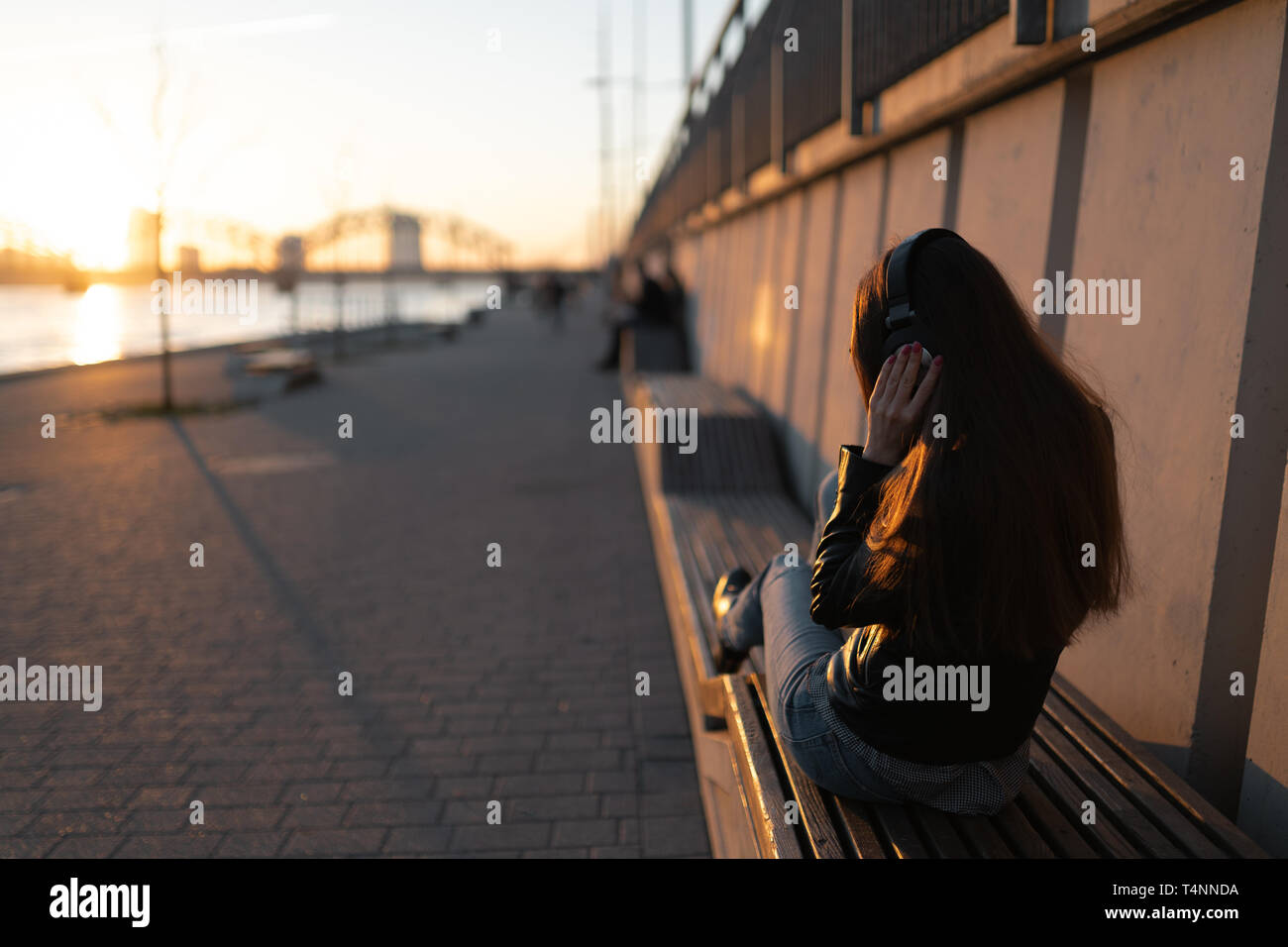 Young woman listens to music in closed headphones through her phone wearing a leather jacket and jeans at a sunset near river Daugava Stock Photo