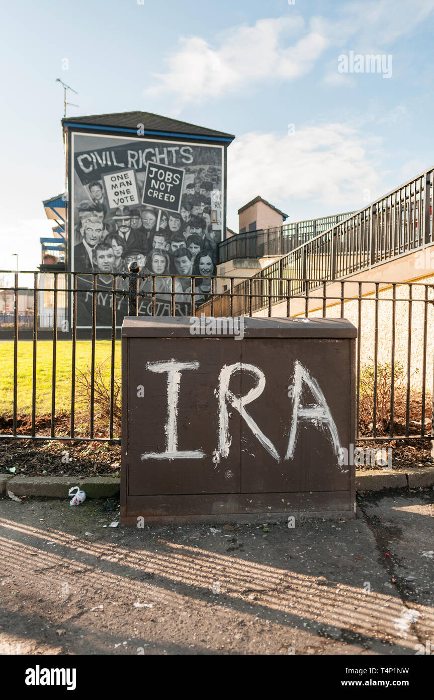 IRA grafittied on a street cabinet with a Republican mural in the background, Londonderry, Derry, Northern Ireland, UK, United Kingdom Stock Photo
