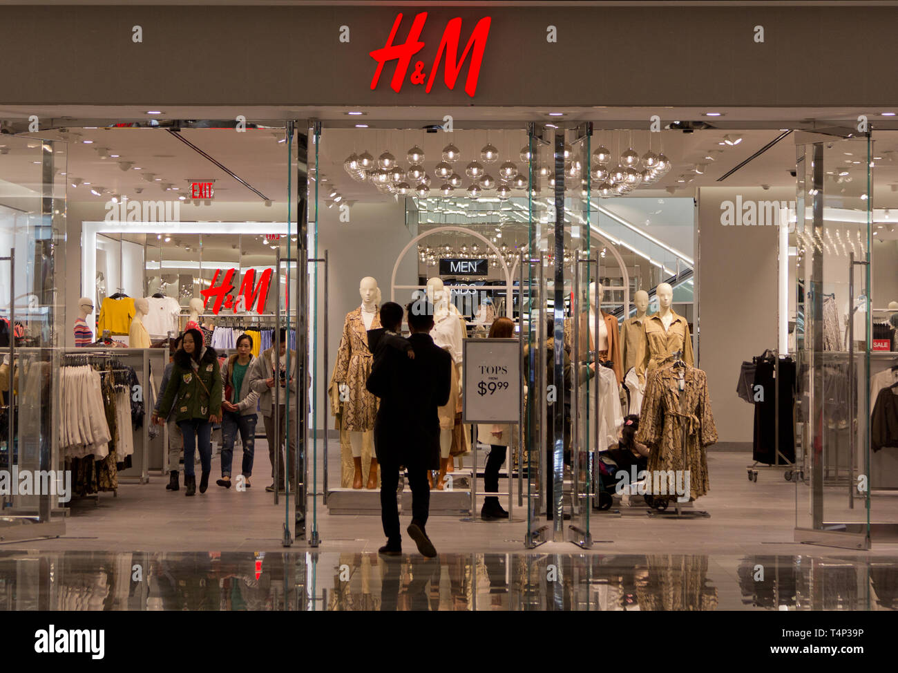 Back view of semi-silhouetted man carrying toddler boy, walking into H&M clothing shop/store at the mall in the new Hudson Yards complex that had opened earlier that month, the highly reflective floor outside the doorway beneath his feet, while shoppers inside browse, or walk toward the exit. Hudson Yards, New York's newest neighborhood, debuted 15 March 2019, to controversy about whether it was too elitist. According to a company press release, the development 'brings together for the first time the West Side and the neighboring districts of Chelsea and Hell’s Kitchen'. Stock Photo