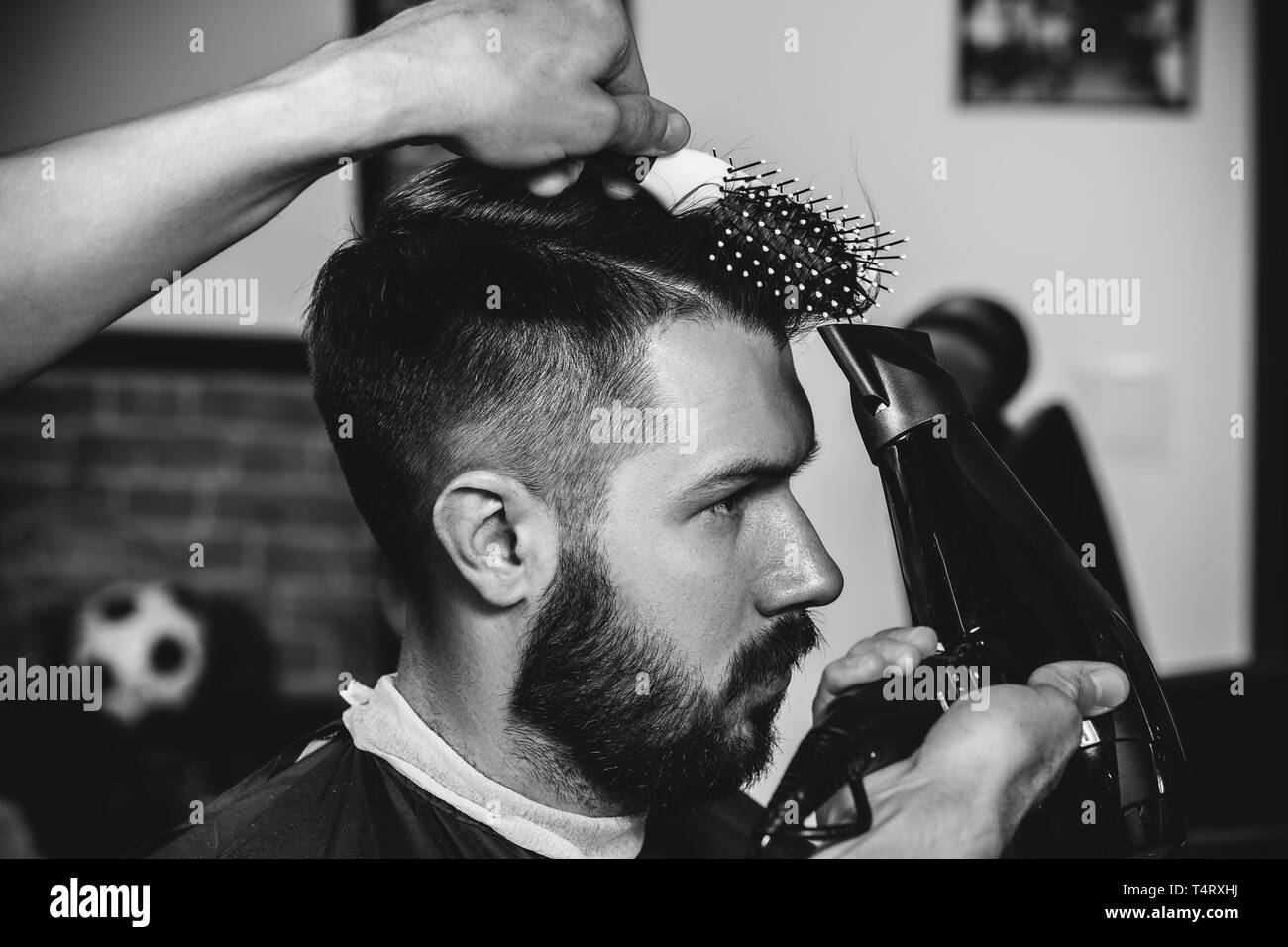 Young handsome barber making haircut for attractive bearded man at barbershop. Black and white or colorless photo. Hairstyle, salon, hairdresser, barber shop, lifestyle concept. Caucasian male models. Stock Photo