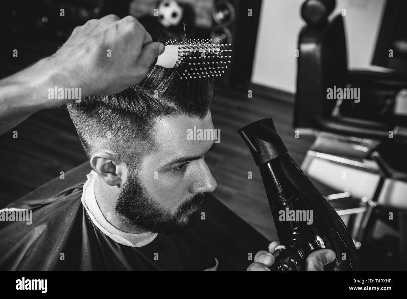 Young handsome barber making haircut for attractive bearded man at barbershop. Black and white or colorless photo. Hairstyle, salon, hairdresser, barber shop, lifestyle concept. Caucasian male models. Stock Photo
