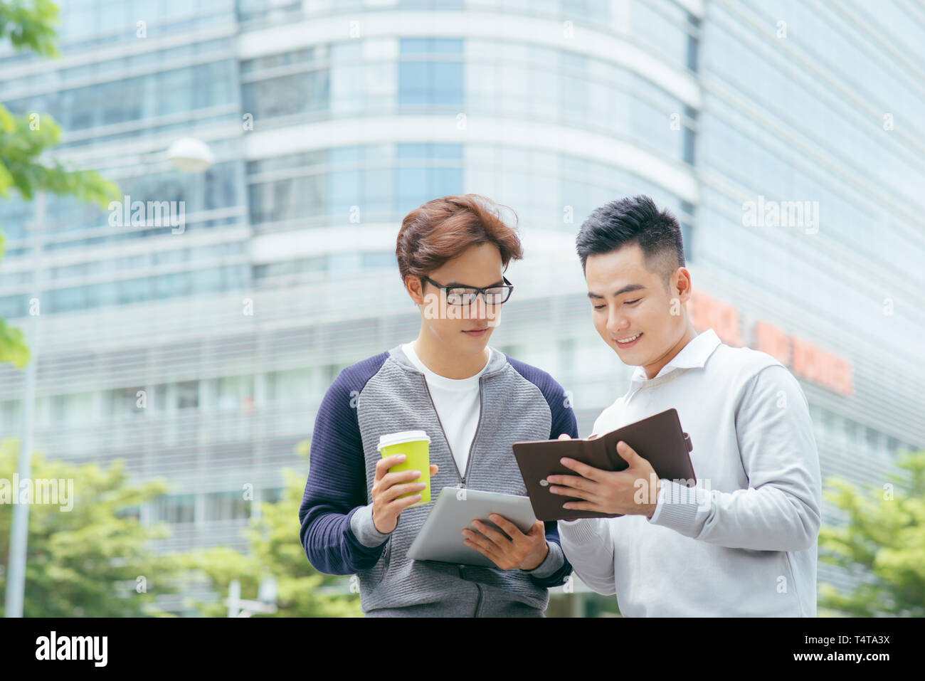 Closeup of two smiling business men using tablet computer and walking with office building in background - Image Stock Photo